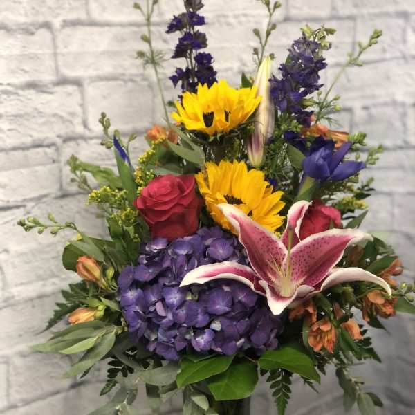 Mixed bouquet with sunflowers, roses, lilies, and purple hydrangeas in a glass vase