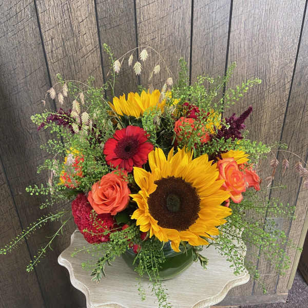 Low arrangement of sunflowers, orange roses, and red blooms in a green bowl on a small table