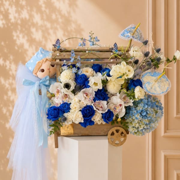Blue and white floral arrangement in a wooden cart with a doll and lace umbrellas