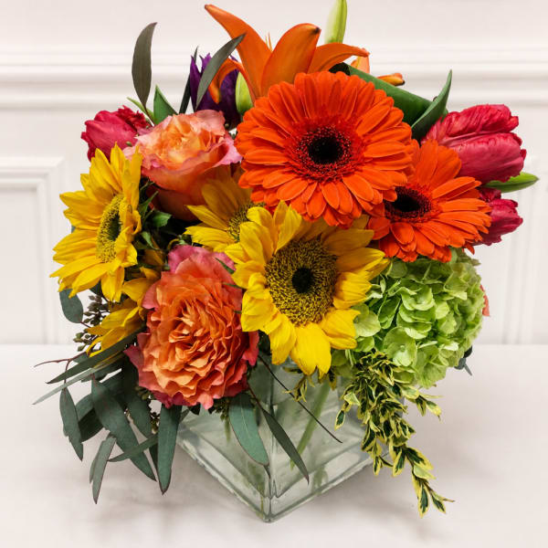 Colorful mixed bouquet in a square glass vase with sunflowers, gerberas, roses, and hydrangea