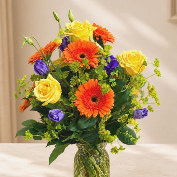 Bouquet of yellow roses, orange gerbera daisies, and purple blooms in a glass vase
