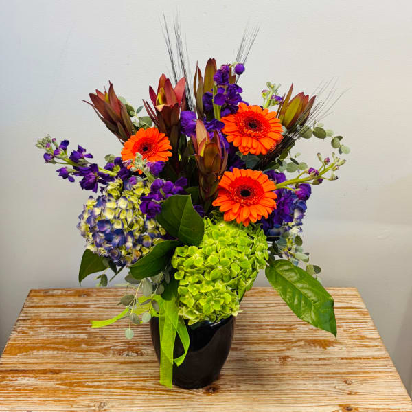 Bright mixed bouquet with orange gerbera daisies, purple blooms, and green hydrangea in a black vase
