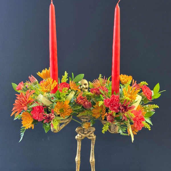 Two red taper candles rise from a colorful floral centerpiece with a skeleton base.