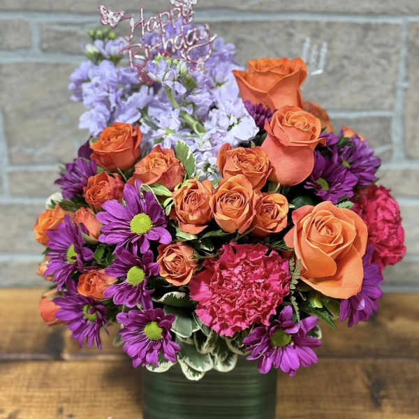 Bright arrangement of orange roses, purple daisies, and pink carnations in a green cube vase with Happy Birthday pick