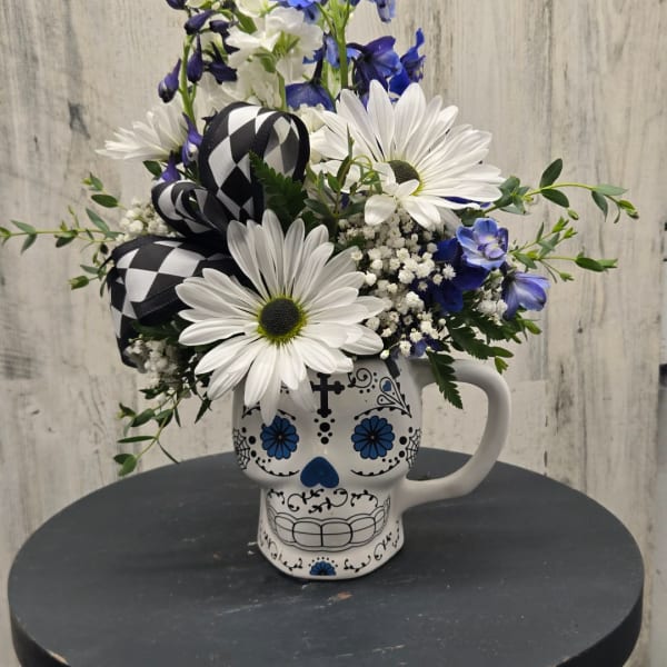 White daisies and blue flowers arranged in a skull mug with a black-and-white ribbon