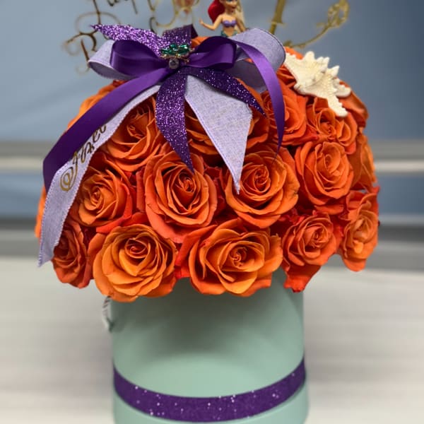 Orange roses arranged in a mint green hatbox with purple ribbon