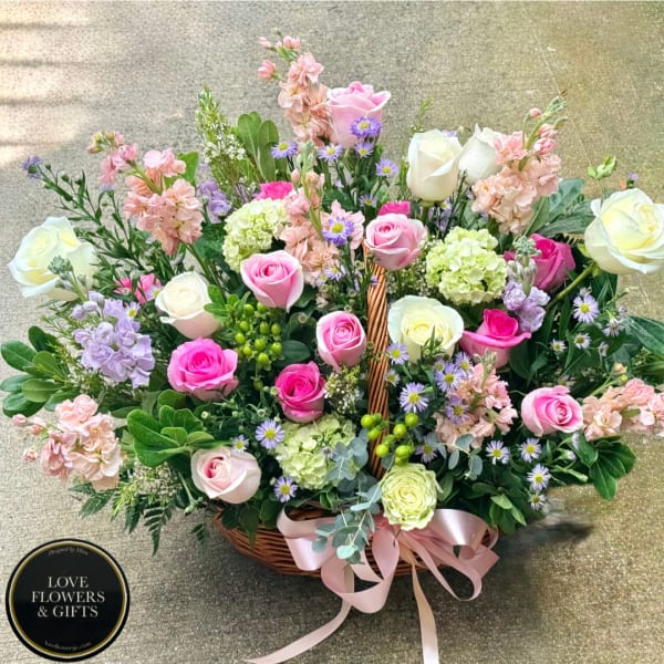 Basket arrangement of pink and white roses with pastel filler flowers and a pink ribbon