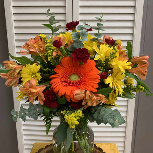 Mixed bouquet with orange gerbera daisies, yellow daisies, and red carnations in a glass vase