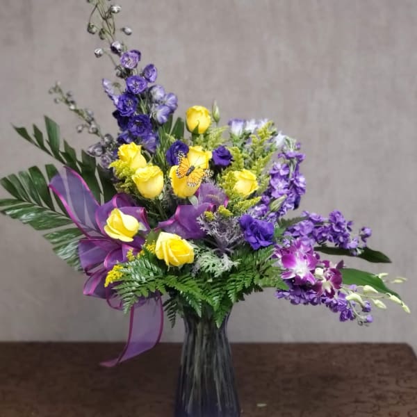 Yellow roses and purple flowers arranged in a glass vase with a ribbon