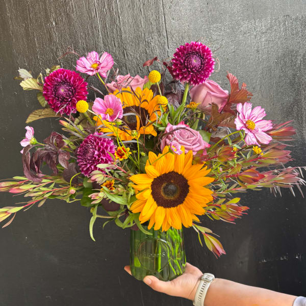 Mixed bouquet with sunflowers, pink roses, and magenta dahlias in a green glass vase