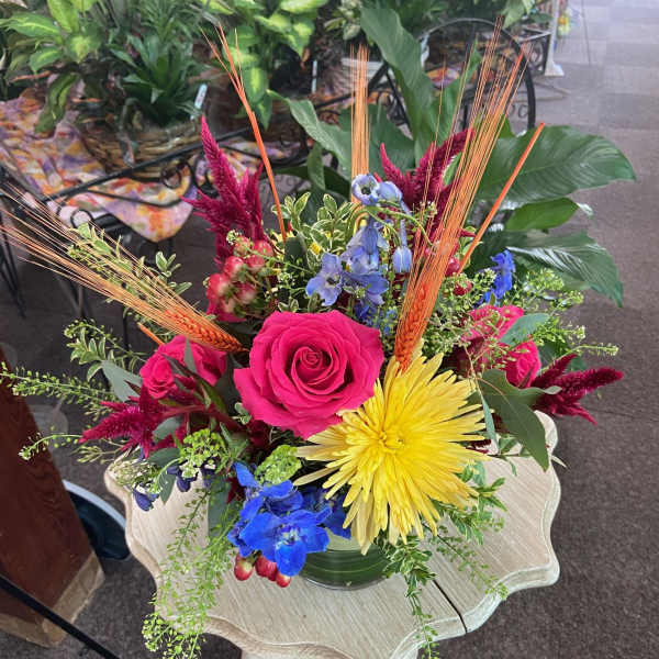Bright mixed arrangement with hot pink rose, yellow mum, blue delphinium and wheat stems in a clear glass vase.
