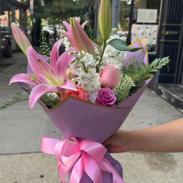 Handheld bouquet of pink lilies, roses, and white flowers wrapped in purple paper