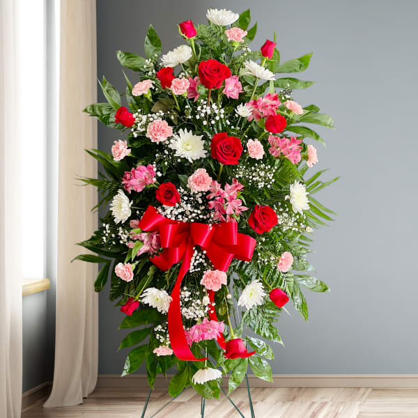 Standing floral spray with red roses, pink carnations, and white daisies on a red ribboned easel