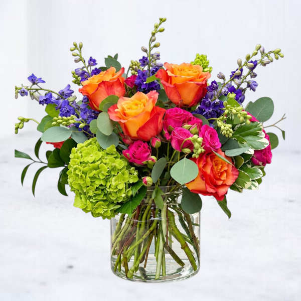 Mixed bouquet of orange roses, pink spray roses, and purple flowers in a glass vase