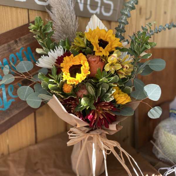 Rustic bouquet of yellow sunflowers, orange roses, and burgundy blooms wrapped in brown paper and raffia.