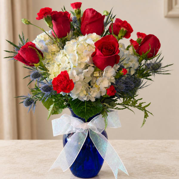 Red roses and carnations arranged with white hydrangeas in a blue vase with a white bow