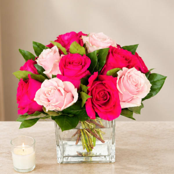 Pink and pale blush roses in a clear square vase beside a small candle