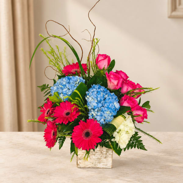 Bright mixed bouquet with pink roses, blue hydrangeas, and gerbera daisies in a square vase.