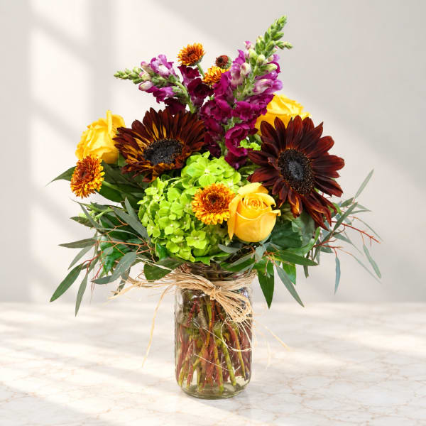 Mixed bouquet of yellow roses, sunflowers, and purple snapdragons in a glass vase