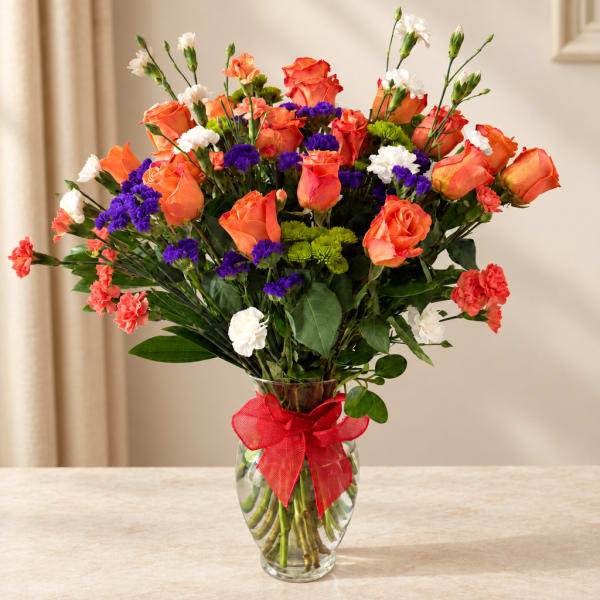Orange roses and purple carnations in a glass vase with a red ribbon
