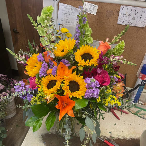 Bright mixed bouquet with sunflowers, lilies, roses, and purple spikes in a glass vase
