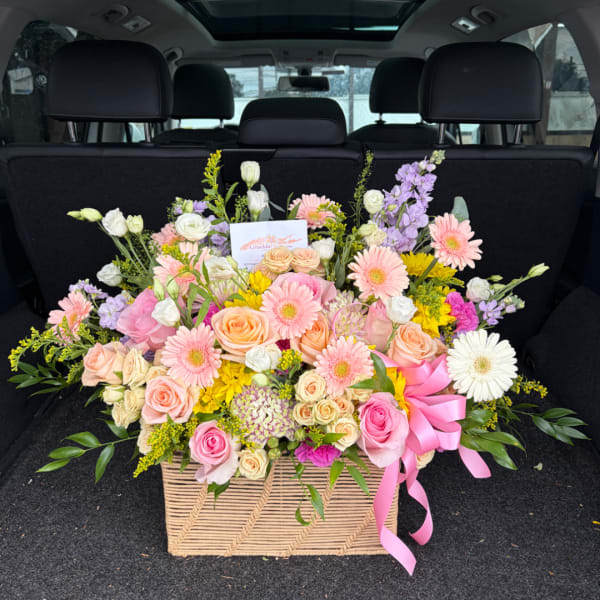 Large mixed flower arrangement in a woven basket with a pink ribbon