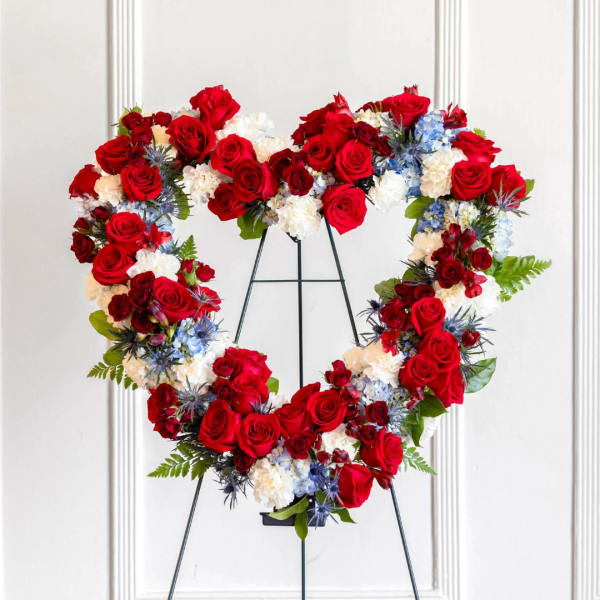 Heart-shaped floral wreath of red roses, white flowers, and blue accents on a stand