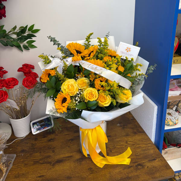 Yellow roses and sunflowers arranged in a white-wrapped bouquet with a yellow ribbon