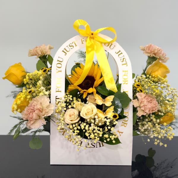 Yellow and peach floral arrangement in a white gift bag with a ribbon handle
