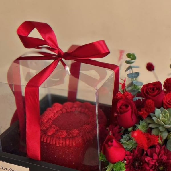 Red cake in a clear box with a red ribbon beside red roses and succulents