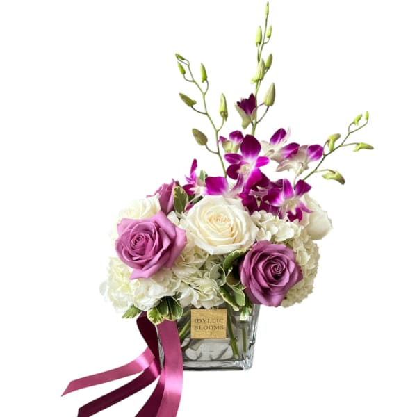Purple roses and white orchids in a clear square vase with a ribbon