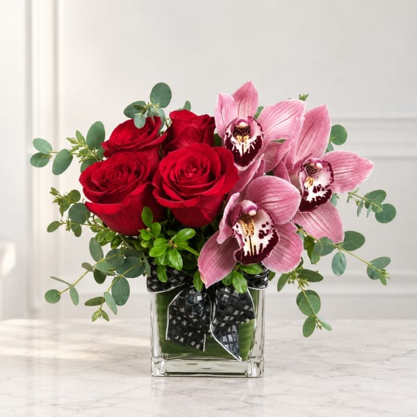 Red roses and pink orchids in a square glass vase with a black ribbon