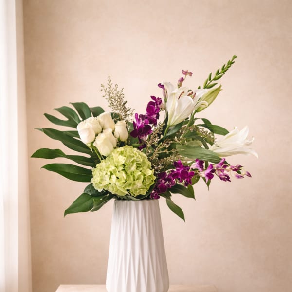 Tall white vase with white roses, lilies, green hydrangea and purple orchids with large tropical leaves.