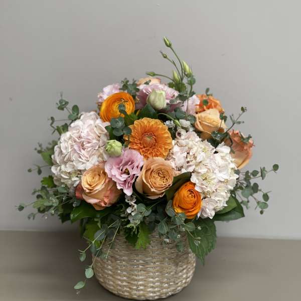 Round arrangement of peach and orange roses, hydrangeas, and pink blooms in a woven basket