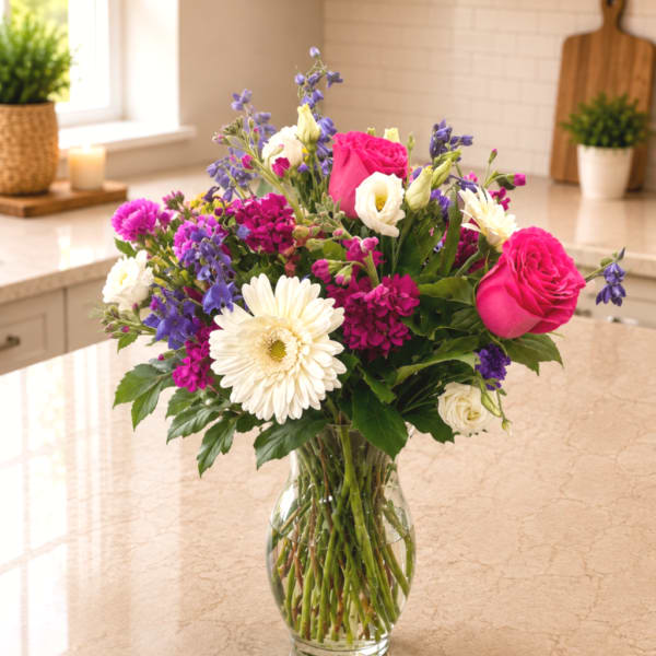 Mixed bouquet of pink roses, white gerbera daisies, and purple flowers in a glass vase