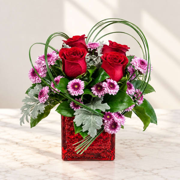 Red roses and pink daisies in a red square vase with looping greenery