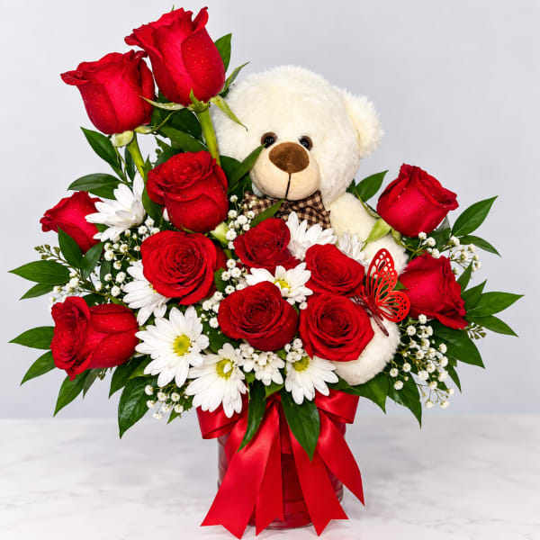 Red roses and white daisies arranged with a teddy bear in a red vase