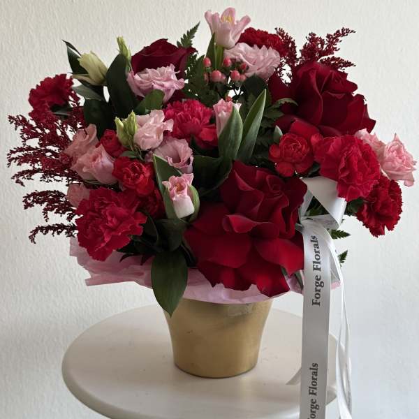 Red and pink mixed flower arrangement in a gold vase with a ribbon
