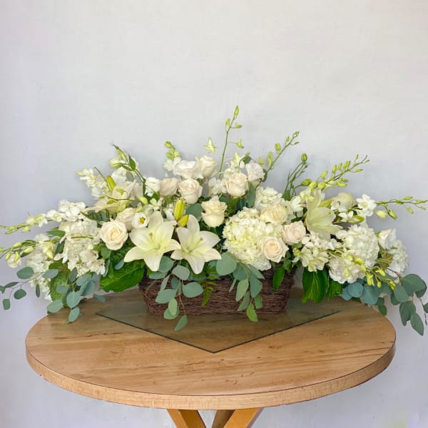 Low rectangular floral arrangement with white lilies, roses, and orchids in a woven basket.