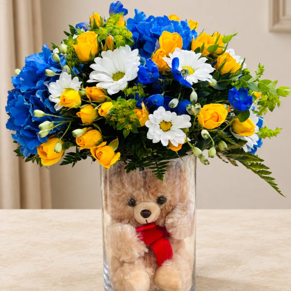 Bouquet of yellow roses, blue carnations, and white daisies in a glass vase with a teddy bear