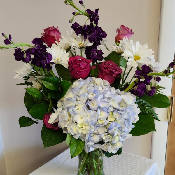 Bouquet with blue hydrangea, pink roses, white daisies, and purple flowers in a glass vase