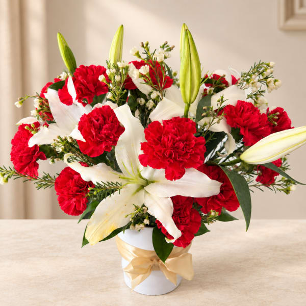 Red carnations and white lilies in a white vase with a gold ribbon