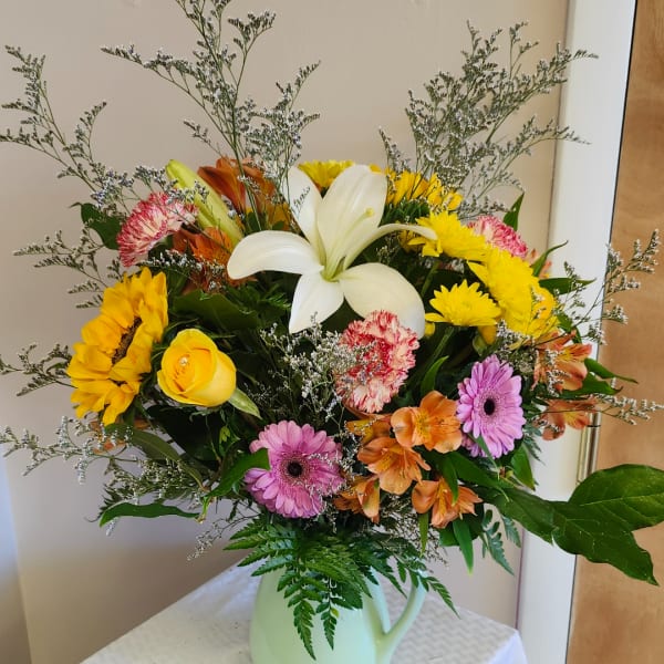 Mixed bouquet in a white pitcher vase with a white lily, sunflowers, gerberas, and carnations