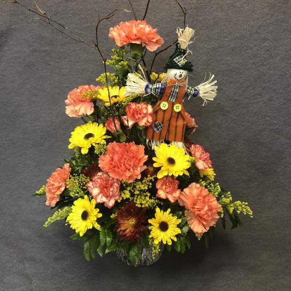 Autumn floral arrangement with yellow daisies and peach carnations, topped with a scarecrow figure.