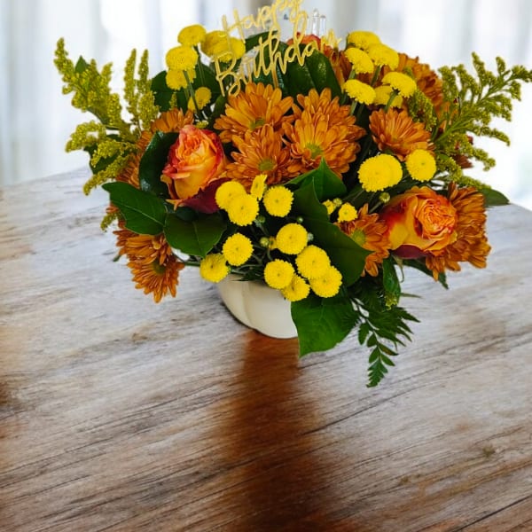 Orange and yellow floral arrangement in a white vase with a birthday topper