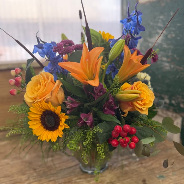 Mixed bouquet with orange lilies, yellow roses, blue delphinium, and a sunflower in a glass vase