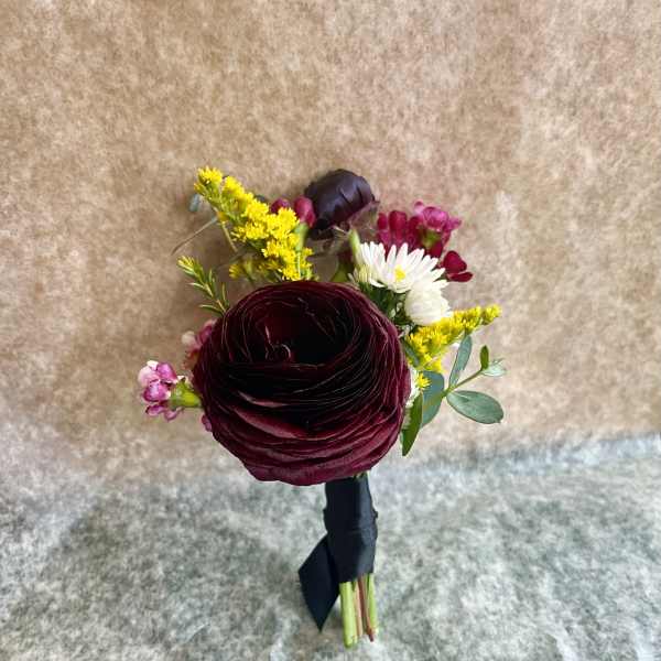 Small boutonniere with dark burgundy ranunculus, yellow and white accents, wrapped with black ribbon