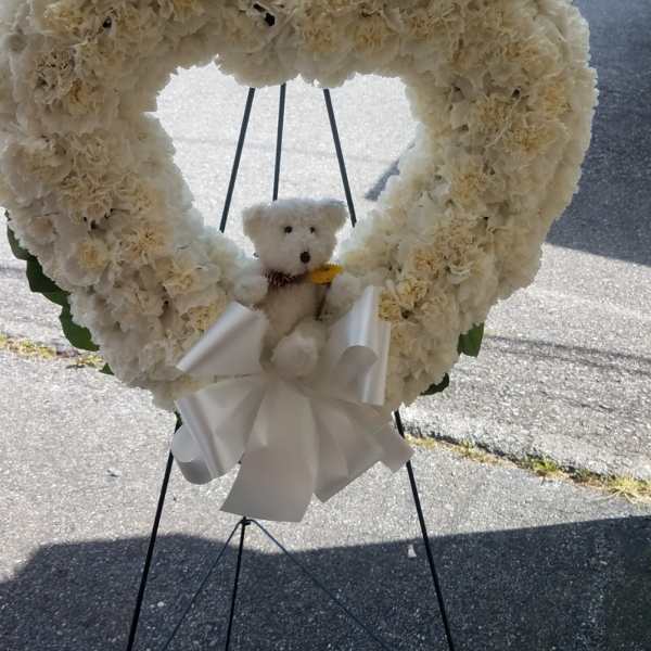 Heart-shaped white floral wreath with a teddy bear and ribbon on a stand
