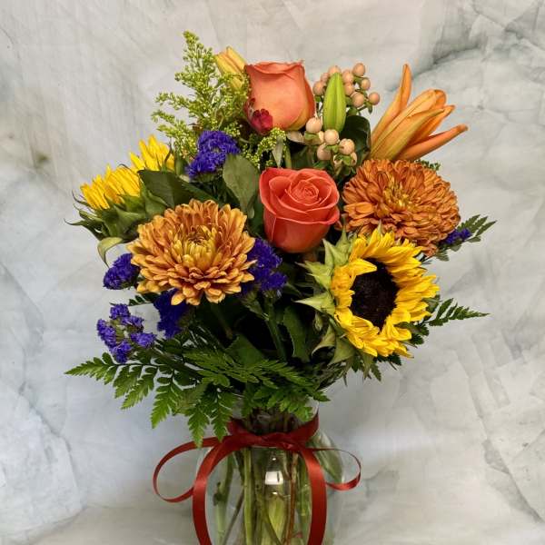 Mixed bouquet of sunflowers, roses, and orange blooms in a glass vase with a red ribbon