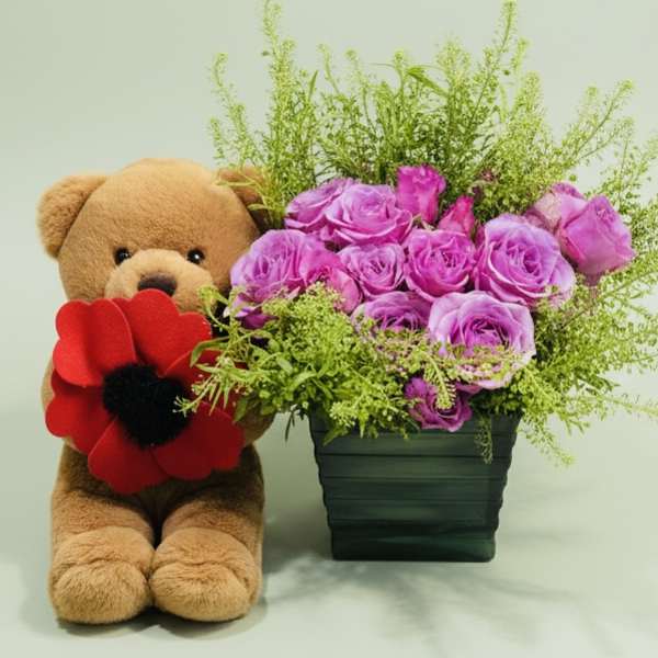 Teddy bear beside a vase of purple roses and greenery